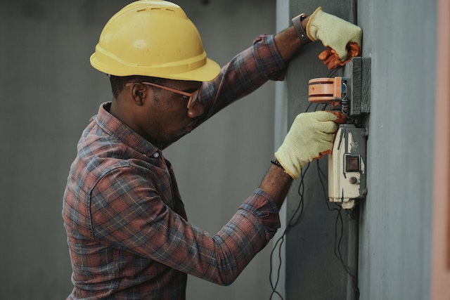 electrician wearing work gloves and a yellow hard hat while inspecting wiring